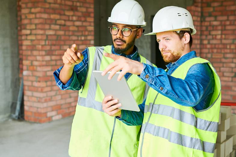 Two construction workers in reflective vests and hard hats discuss plans while looking at a tablet. They are standing indoors near a brick wall, and one is pointing in the distance.