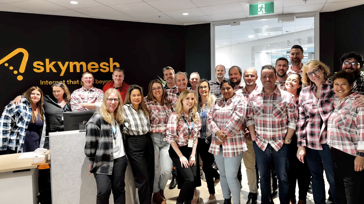 A group of smiling people, many wearing red and white plaid shirts, pose together in an office lobby in front of a black wall with the yellow Skymesh logo and slogan.