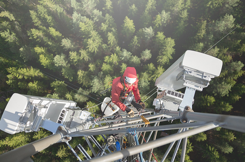 A person in safety gear climbs a tall communication tower surrounded by a dense forest. The image is taken from above, showing various equipment on the tower and a clear view of the treetops below.