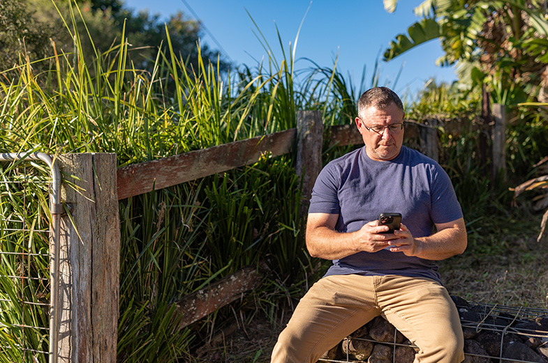 A man in a blue shirt and khaki pants sits on a stone bench outside, checking his phone. He's near a rustic wooden fence with tall grass and a bright blue sky in the background.