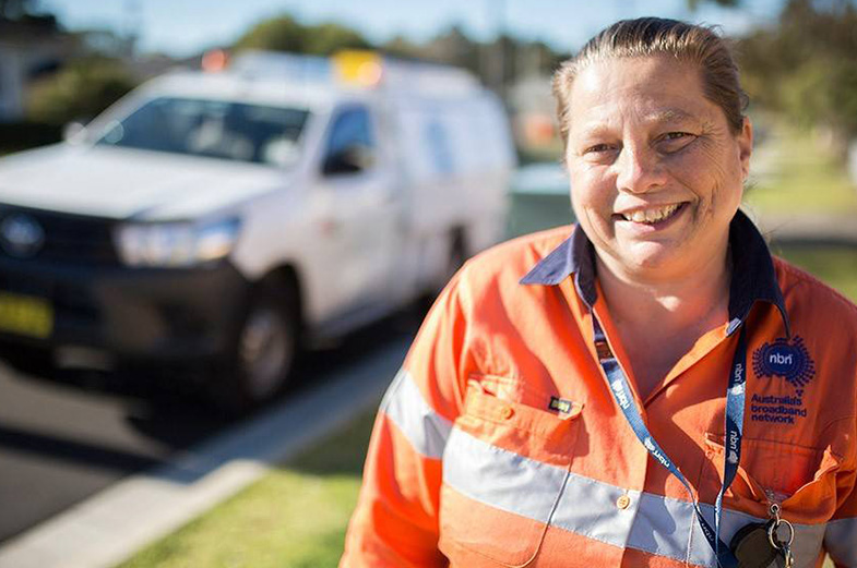 A person in an orange high-visibility jacket smiles at the camera while standing outdoors. A white utility vehicle with blurred text is parked on the road in the background.