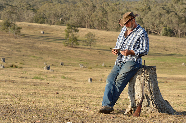 A man in a plaid shirt and cowboy hat leans against a tree stump in a grassy field, looking at a phone. A rifle rests against the stump. The background features scattered trees and open land.