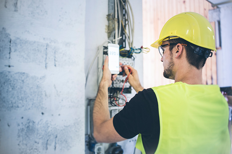 A construction worker wearing a yellow hard hat and vest is using a multimeter to check voltage in a circuit breaker box. The worker is inside a building, next to a white brick wall.