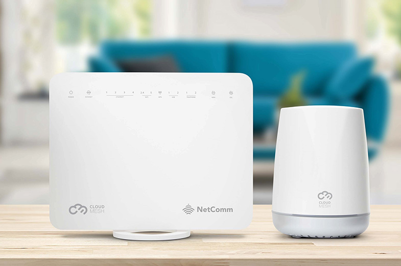A white Cloud Mesh Wi-Fi router and matching satellite device are placed on a wooden table. A blue couch with cushions and a window with light coming in is blurred in the background.