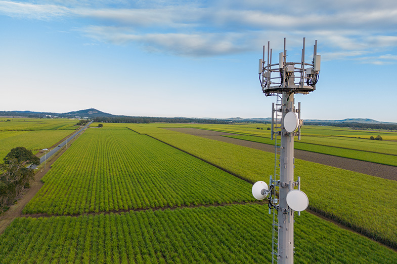 Aerial view of a telecommunications tower with various antennas situated in a vast, green agricultural landscape. Fields extend to the horizon with a clear blue sky overhead.