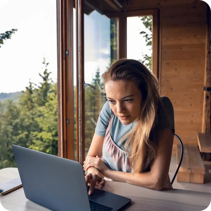 A woman with long hair sits at a table using a laptop in a wooden room with large open windows, overlooking a forested landscape with trees and hills in the background.